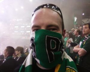 A person is at a Timbers game, with his sunglasses on his head and a Timbers scarf covering his nose and mouth.