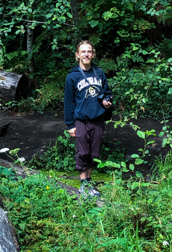 A person stands smiling in nature and looking at the calendar, he wears a Columbia sweatshirt.