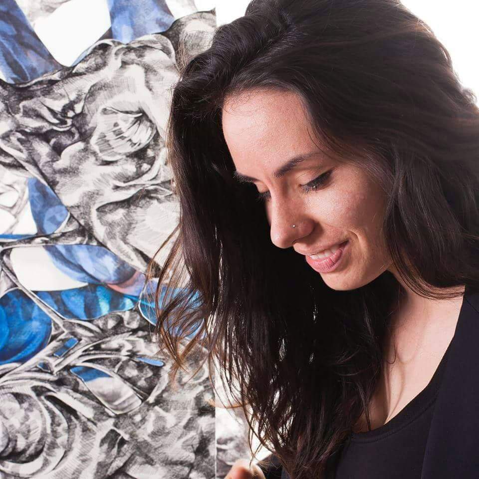 A woman with long flowing hair that cascades over her shoulder looks down and smiles in front of a blue and grey work of art.