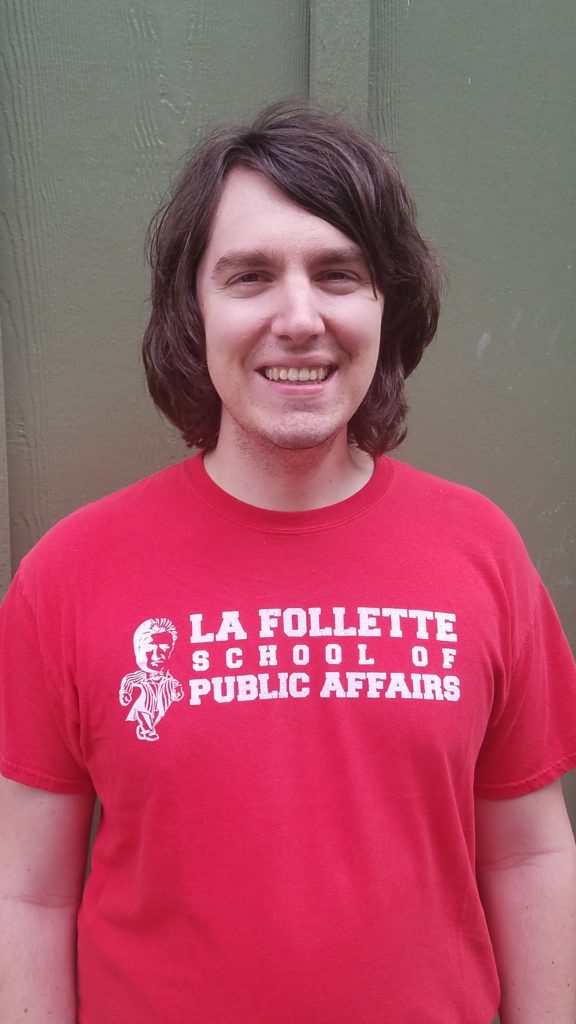A smiling man with a shaggy haircut stands in front of a wall. He wears a red t-shirt that reads La Follette School of Public Affairs. 